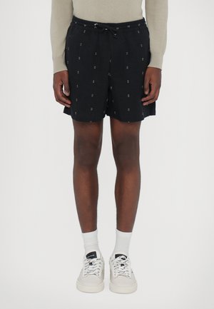 Schwarze Shorts mit einem elastischen Bund und Kordelzug, die ein dezentes geometrisches Muster in Weiß aufweisen. Kombiniert mit weißen Socken und Turnschuhen.