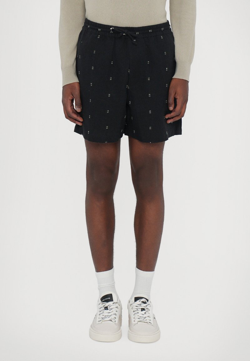 Schwarze Shorts mit einem elastischen Bund und Kordelzug, die ein dezentes geometrisches Muster in Weiß aufweisen. Kombiniert mit weißen Socken und Turnschuhen.