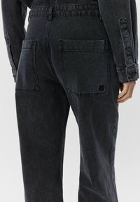 Sötét farmer kezeslábas, laza fit, dupla hátsó zsebekkel és texturált anyaggal. Részlet a fémből készült snap zárakról és varrásról.