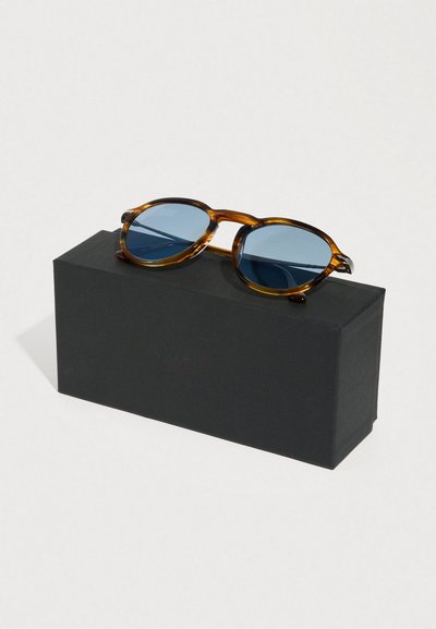 Occhiali da sole tartarugati con lenti blu, montatura rotonda e accenti marrone scuro, appoggiati su una scatola rettangolare nera opaca.