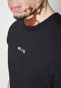 Musta t-paita pyöreällä kaula-aukolla, jossa lukee "AIR" valkoisilla ja vihreillä kirjaimilla. Sujuva rakenne, klassinen istuvuus. Huomattava ihon väripigmentaatiomalli lähellä.