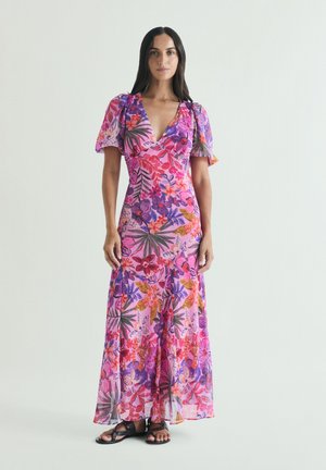 Femme aux longs cheveux foncés portant une robe maxi fleurie colorée avec des manches bouffantes et des sandales noires, debout devant un fond uni.