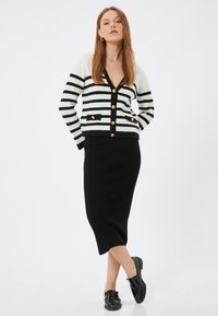 Gestreifter schwarz-weißer Cardigan mit goldenen Knöpfen, kombiniert mit einem schwarzen, gerippten Midirock und schwarzen Schuhen, vor einem schlichten Hintergrund.