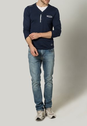 Navy long-sleeve shirt with a V-neck, featuring a white button placket and a chest pocket. Paired with light blue jeans and sneakers.