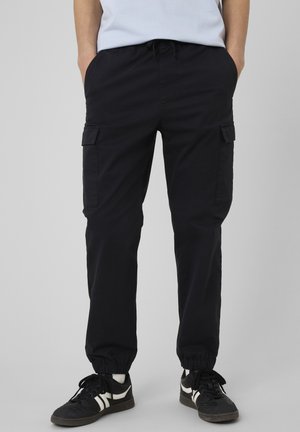 Personne portant un pantalon cargo noir avec des poignets élastiques et des baskets noires avec des bandes blanches, debout les mains dans les poches.