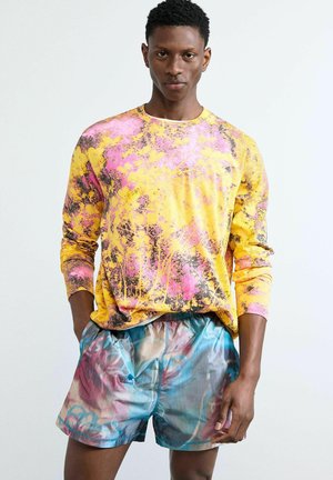 Hombre con camisa de manga larga salpicada de amarillo y rosa y pantalones cortos con patrón abstracto multicolor, de pie contra un fondo liso.