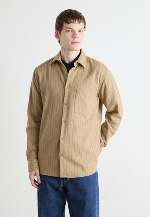 Beige Hemd mit vertikalen blauen Streifen, ausgestattet mit einer Brusttasche und einem lässigen Schnitt, kombiniert mit blauen Denim-Hosen.