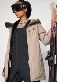 Beige skijakke med hette, svart skibukse, mønstret topp. Modellen holder ski med bindinger. Har logo-patcher og glidelåser.