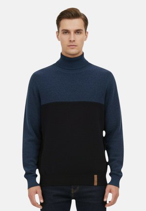 Giovane uomo con indosso un maglione a collo alto a blocchi di colore blu e nero e jeans scuri, in piedi davanti a uno sfondo chiaro e uniforme.