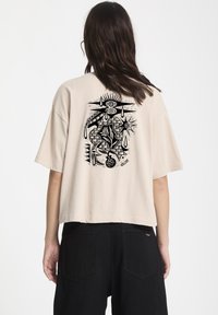 Beige lyhyempi t-paita, jossa on musta graafinen kuvio selkäpuolella, sisältäen silmiä, auringon ja abstrakteja kuvioita. Lyhyet hihat, rento istuvuus. Mustat housut näkyvissä.