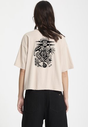 Beige cropped t-shirt met zwart grafisch ontwerp op de achterkant, met ogen, zon en abstracte patronen. Korte mouwen, relaxte pasvorm. Zwarte broek zichtbaar.