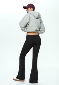 Gray cropped hoodie, black flared pants, back view. Model wears a maroon cap and white shoes with a light brown sole. Smooth textures, casual design.