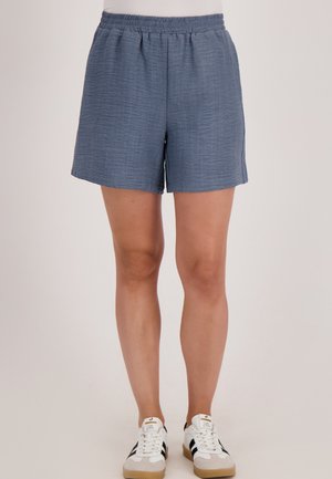 Blauwe textuurshorts met een elastische tailleband, gedragen door een persoon die staat, gecombineerd met witte sneakers met zwarte strepen.