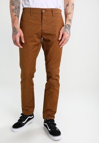 Bruine chino's met een gladde textuur, slim fit en zichtbare voorste knoop. Gekoppeld aan zwarte sneakers met witte accenten.