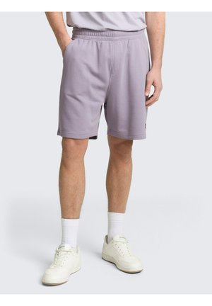 Man draagt lichtpaarse elastische shorts met zijzakken, witte sportsokken en witte sneakers, staand tegen een effen achtergrond.