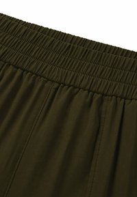 Olijfgroene broek met elastische tailleband en een zachte textuur, voorzien van horizontale plooien langs de tailleband en verticale naden op de benen.