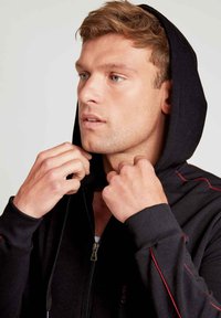 Zwarte zip-up hoodie met rode accentstiksels, voorzien van een trekkoordcapuchon en geribde mouwen. Model past de kraag met de handen aan.