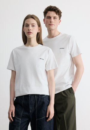 Due giovani adulti stanno fianco a fianco indossando t-shirt bianche con il logo ARKK; donna con jeans scuri, uomo con pantaloni scuri a righe sottili.