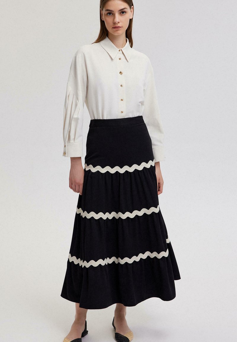 Chemise blanche à manches bouffantes avec des boutons dorés, associée à une jupe noire à volants avec des accents crème ondulés. Texture de tissu lisse.