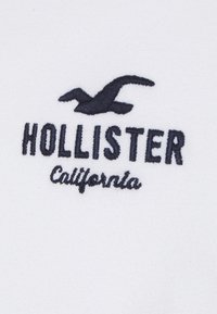 Weißer Stoff mit marineblauem gesticktem Text, der "HOLLISTER" und "California" liest, sowie einer Silhouette einer Möwe.