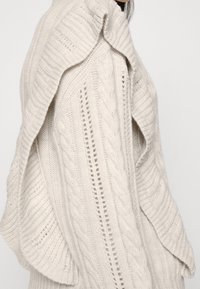 Gros plan sur un pull crème en maille torsadée avec des textures côtelées et à motifs, présentant des manches larges et drapées sur une personne.