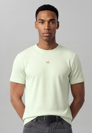 Νέος άνδρας που φοράει ένα ανοιχτοπράσινο ριμπ μπλουζάκι με μικρό πολύχρωμο λογότυπο "CK", στέκεται μπροστά σε απλό γκρίζο φόντο.