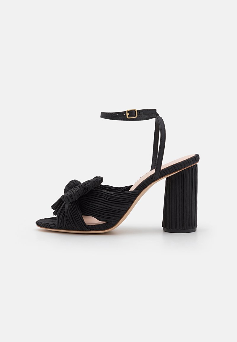 Sandalia de tacón negro con un upper de tela texturada y plisada. Presenta un diseño anudado en la parte frontal y un tacón grueso. Correa en el tobillo ajustable.