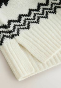 Uldtrøje i creme med sort zigzag-mønster på brystet. Har ribbede cuffe og kant, hvilket giver en blød, tekstureret finish.