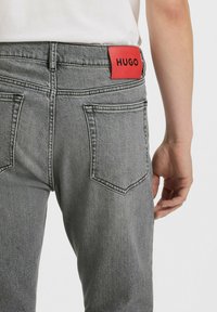 Grå denim jeans med baglommer, forsynet med en rød rektangulær label med teksten "HUGO" på taljen, vist fra siden.