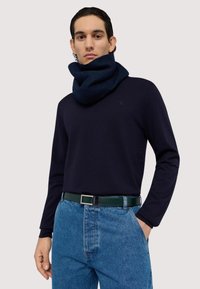 Marineblaues Langarmoberteil mit einem gerollten Schal, kombiniert mit hochgeschnittenen blauen Jeans und einem grünen Gürtel mit rechteckiger Schnalle.