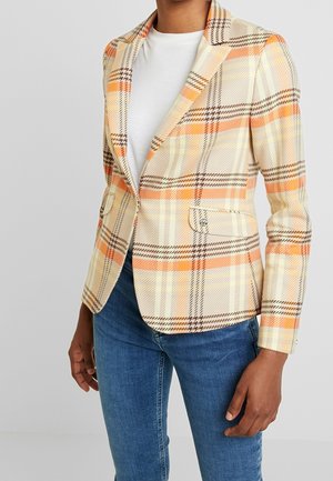 Mujer con blazer entallado de cuadros beige, naranja y marrón, sobre una camisa blanca y jeans azules, de pie con una mano relajada.