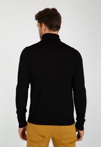 Maglione a collo alto nero realizzato in tessuto a maglia, con maniche lunghe e polsini a coste, visto da dietro, abbinato a pantaloni khaki.