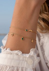 Goldene Armbandketten mit grünen Steinen, in verschiedenen Formen wie Kreisen und Halbmonde, vor einem lichttexturierten Ärmel.