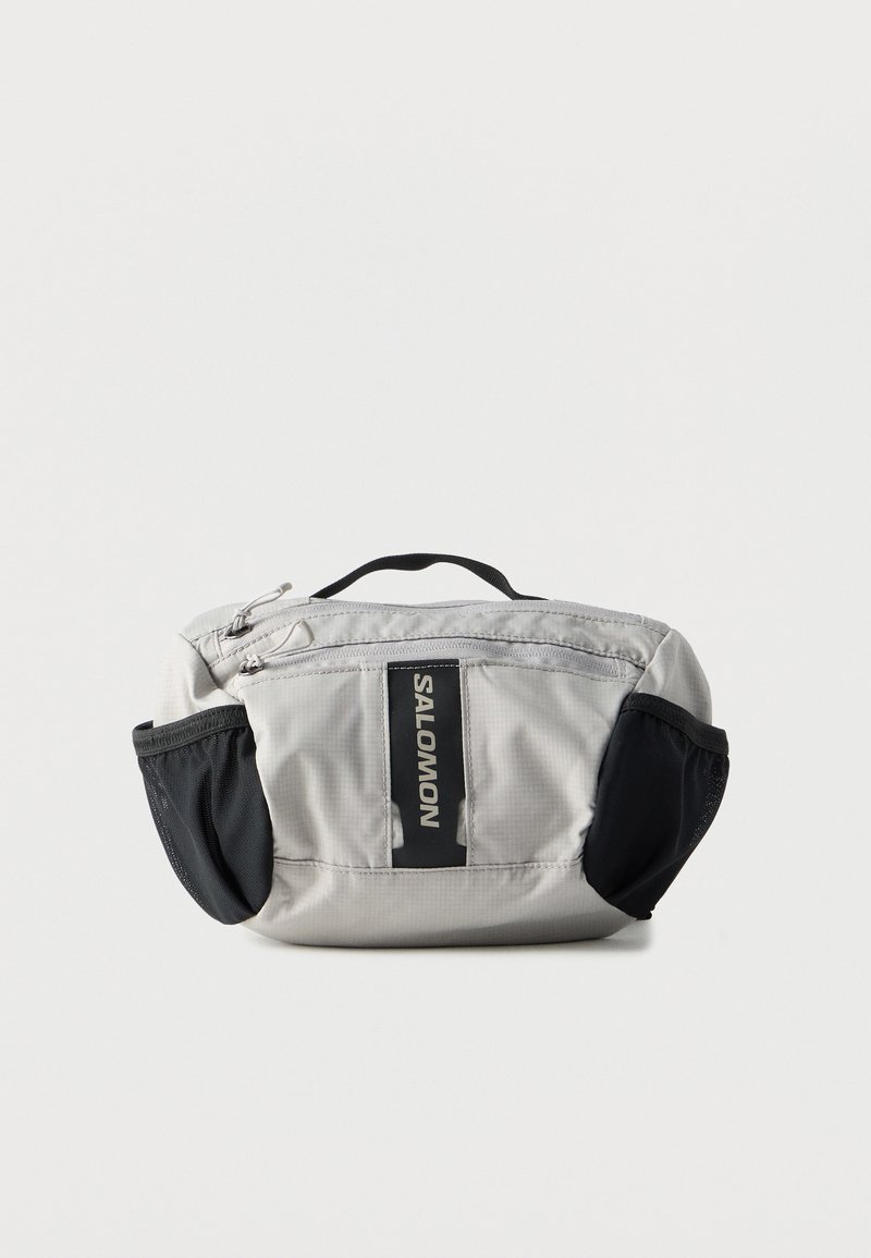 Pochette en tissu gris avec des côtés en mesh noir, dotée d'une poignée supérieure et d'une fermeture éclair. Présente un logo noir bien visible sur le devant.