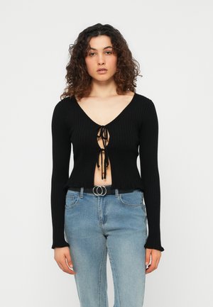 Mujer con cabello rizado que lleva una blusa negra de manga larga con tejido acanalado y anudada al frente, combinada con jeans azul claro y un cinturón de doble hebilla.