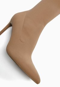 Vaaleanruskea polven korkeudelle ulottuva teräväkärkinen saapas, jossa on sileä, teksturoitu pinta sekä saagan reunoilla ja sivuilla näkyvät saumalävikkeet.