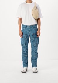 Pantalones cargo azul claro con múltiples bolsillos, un ajuste relajado y una textura desgastada, combinados con una camiseta blanca holgada y zapatillas blancas.