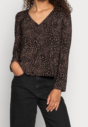 Vrouw draagt een blouse met lange mouwen, V-hals, bruin en zwart dierenprint en een zwarte jeans, één hand in de zak, kijkt vooruit.