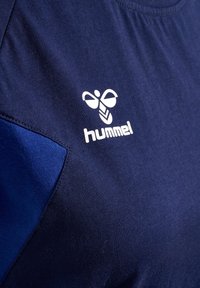T-paita tummansinisellä värillä, lyhyet hihat, jossa on teksturoitu kangas ja valkoinen "hummel"-logo sekä suunnitteluelementti vasemmassa rinnassa.