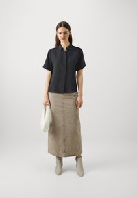 Sort kortærmet skjorte med knapper samlet med en knælang beige denimnederdel. Modellen holder en hvid håndtaske og har lysegrå ankelstøvler på.