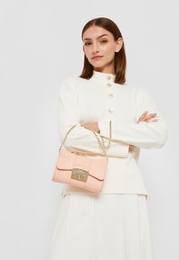 Růžová kožená kabelka se zlatým řetízkem, obdélníkový tvar, klopové zapínání a dekorativní kovové prvky. Model nosí krémový top s knoflíky.