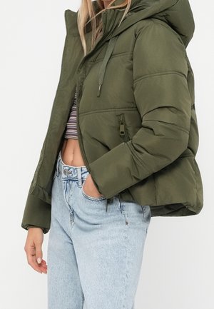 Femme portant une veste matelassée vert olive avec capuche et un jean taille haute bleu clair, main dans la poche, top court rayé visible en dessous.