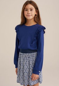 Modra dolga majica z rebrasto teksturo in naborki na ramenih, v kombinaciji z vzorčasto krilo z modro-belim cvetličnim vzorcem.