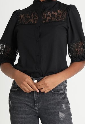 Kobieta w czarnej bluzce zapinanej na guziki z koronkowymi panelami na dekolcie i rękawach, zestawiona z wysokim stanem podartymi ciemnoszarymi dżinsami.