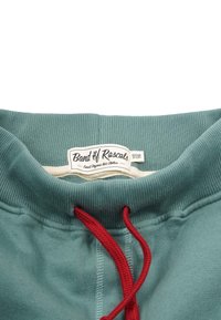 Ekologisk bomullströja med krage i teal och ribbad textur, med en röd dragsko och en vit etikett som visar varumärkets namn och skötselråd.