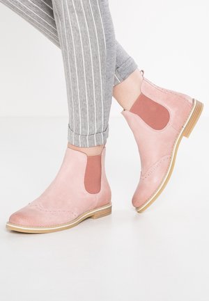 Pink suede ankle boots with elastic side panels, decorative stitching, and a light brown sole. Worn with gray striped pants.