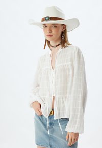 Weißes, strukturiertes Hemd mit langen Ärmeln und Kordelzug in der Taille, kombiniert mit einem Jeansrock; ergänzt durch einen breitkrempigen Hut und einen Choker.