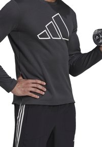 Schwarzes langärmeliges Sportshirt mit weißem Adidas-Logo, strukturierter Stoff, kombiniert mit schwarzen Hosen mit weißen Seitenstreifen.