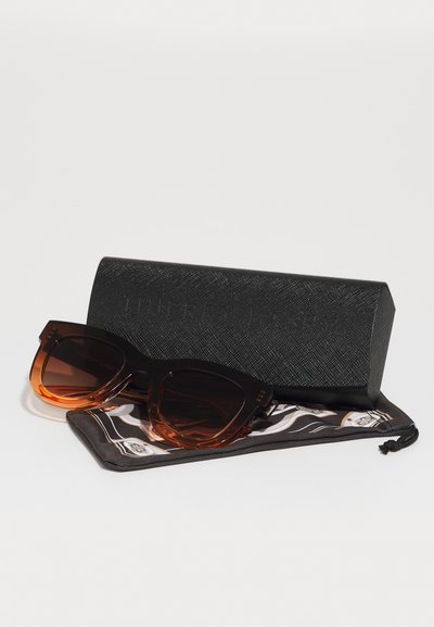 Ochelari de soare cu lentile de nuanță brună gradient, ramă neagră și cu imprimeu țestoasă, așezați pe o țesătură cu model, lângă o husă texturată neagră.