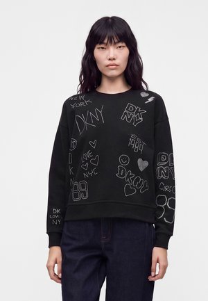 Jeune femme portant un sweat-shirt noir avec un texte blanc et des dessins de cœurs, debout devant un fond clair uni.